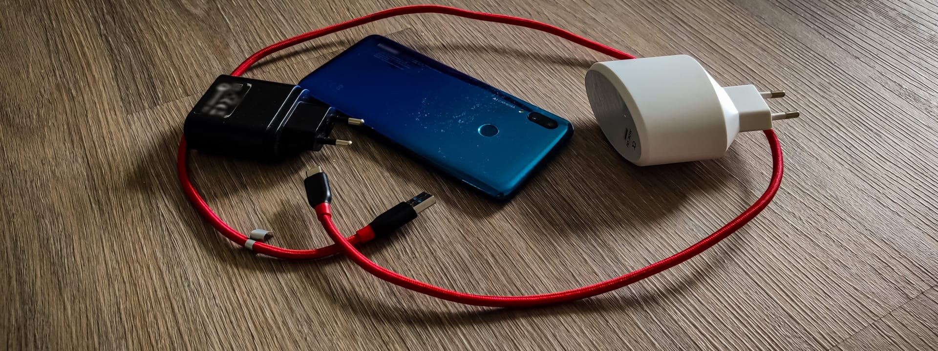 Auf dem Bild sind ein blaues Smartphone, ein rotes Ladekabel und zwei verschiedene Netzteile auf einem Holzfußboden zu sehen.
