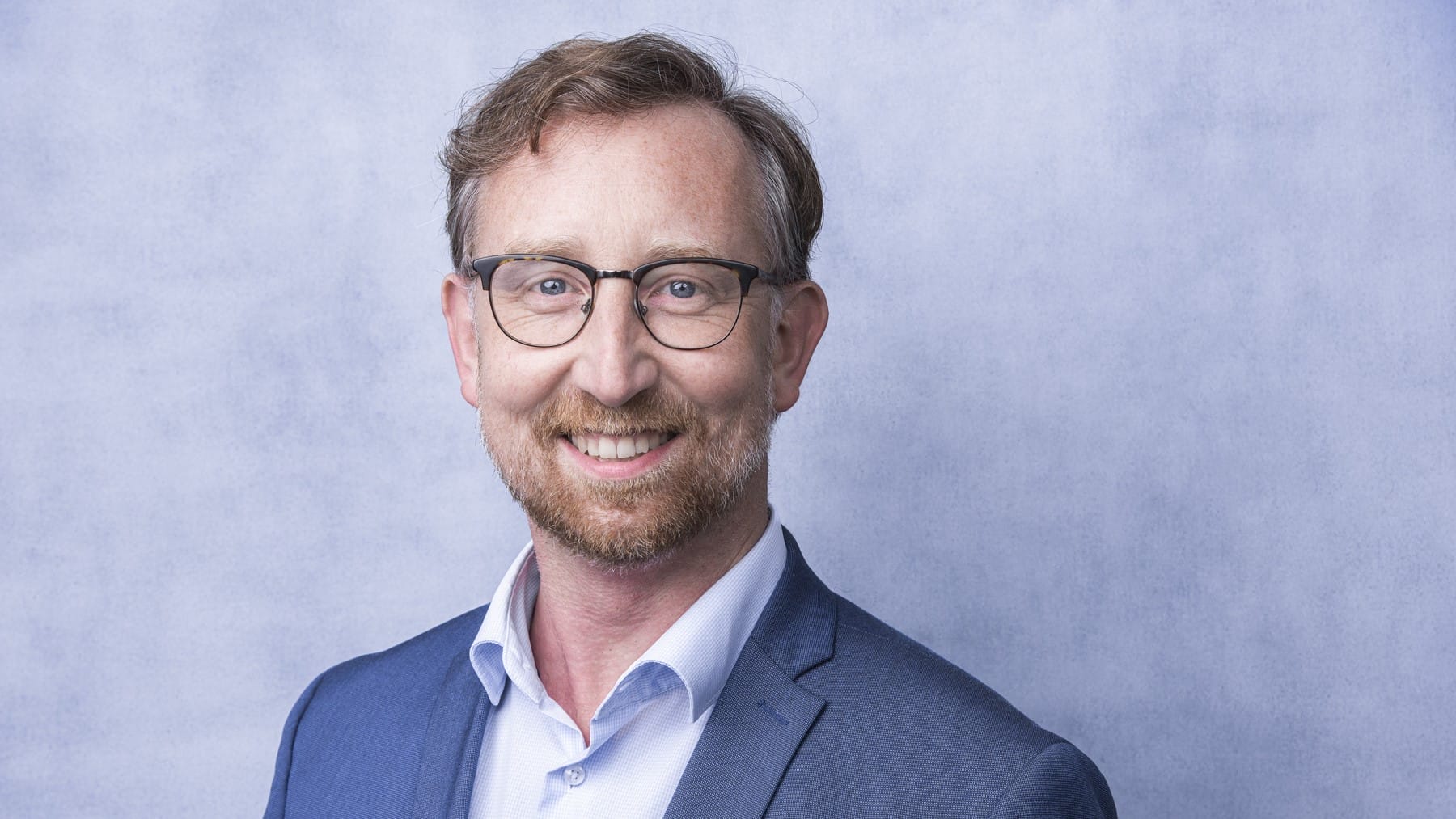 Portrait: Mann im Anzug lächelt in eine Kamera vor einem grauen Hintergrund