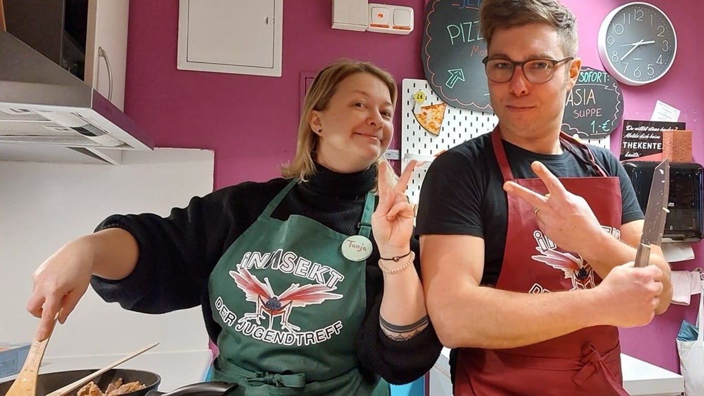 Foto: Tanja und Daniel vom iNNSEKT Team posen in der Küche des Jugendtreffs: Tanja rührt in einer Pfanne, Daniel hat Zwiebeln klein geschnitten