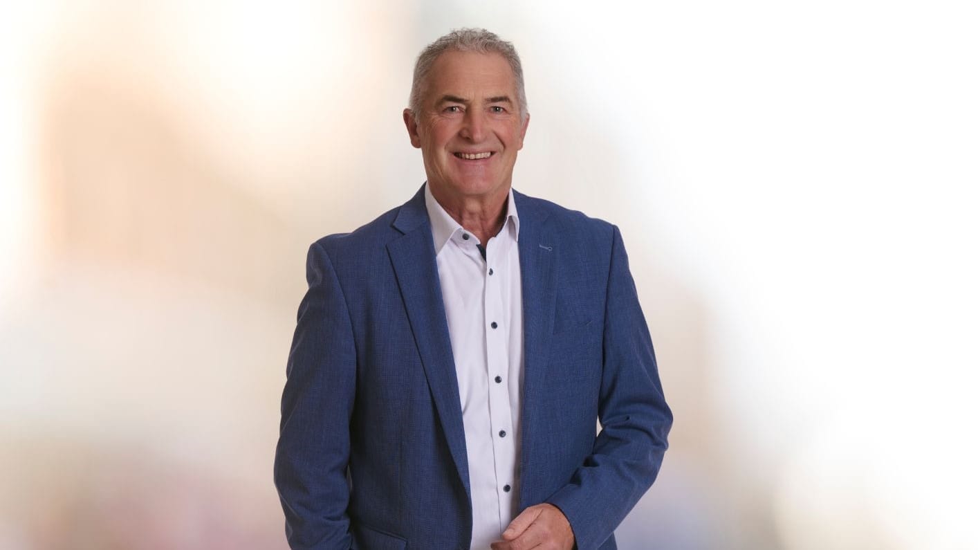 Portrait: Mann mit Anzug steht vor unscharfem Hintergrund und lächelt