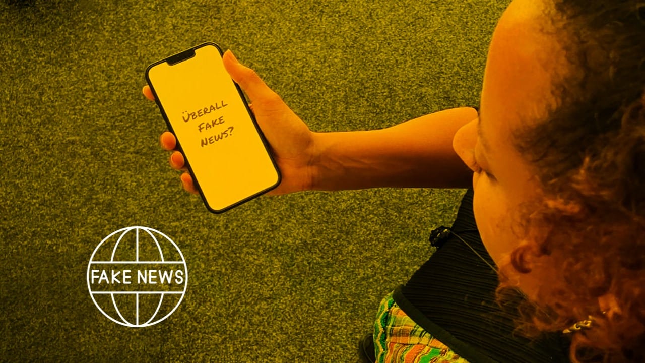 Foto: Eine junge Frau schaut auf ihr Handy, worauf die Frage: "Überall Fake News?" zu lesen ist.