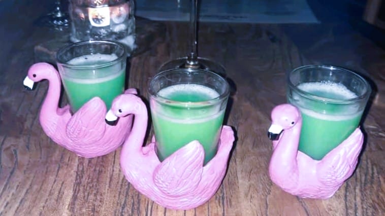 Foto: Auf einem Tisch stehen drei Gläser, gefüllt mit einer grünen Flüssigkeit, die in rosafarbenen Halterungen in Form von Schwänen stecken