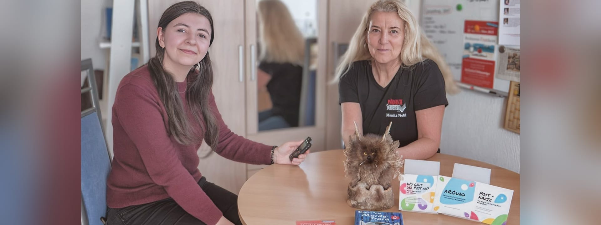 Foto: Zwei Frauen sitzen an einem runden Tisch, die Frau links hält ein Aufnahmegerät in der Hand. Beide lächeln in die Kamera