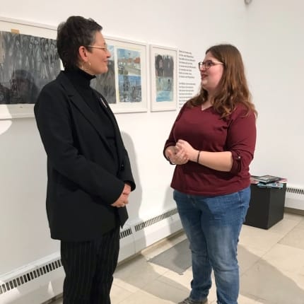 Foto: Zwei Menschen im Gespräch vor gerahmten Bildern und Buch-Auszügen an der Wand