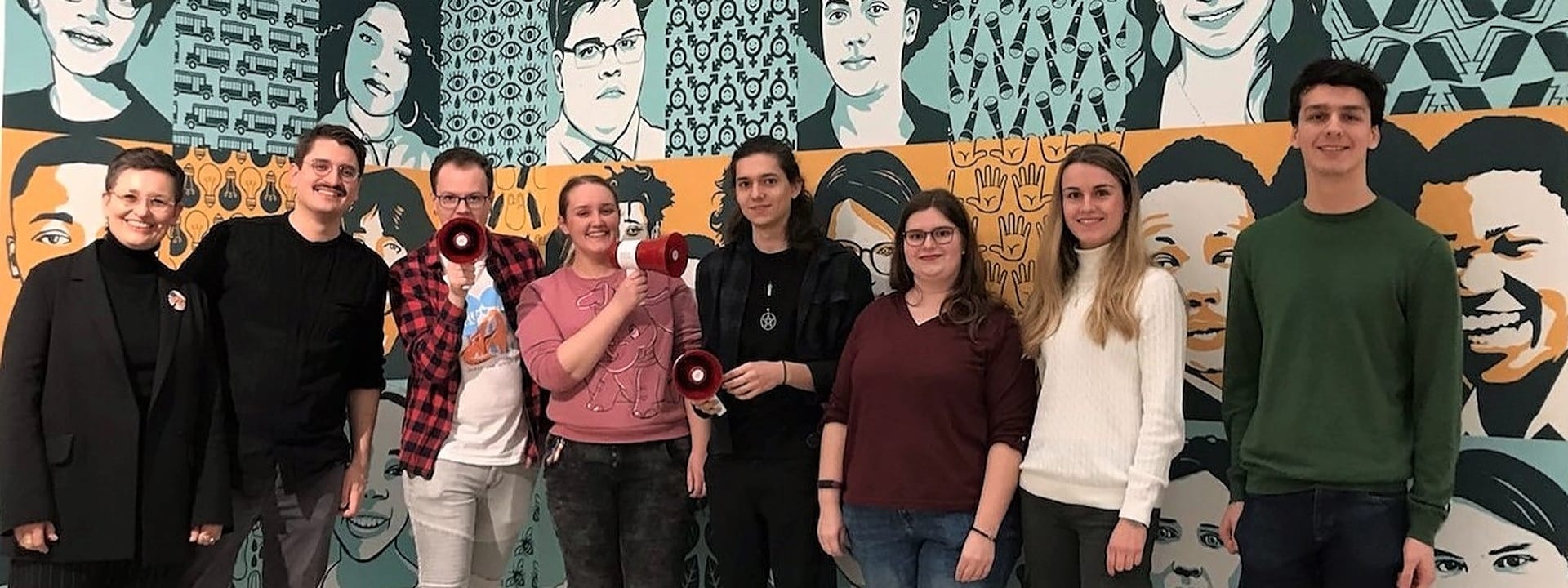 Foto: Acht Personen stehen vor einer Comic-Wand, drei davon haben ein Megafon in der Hand.