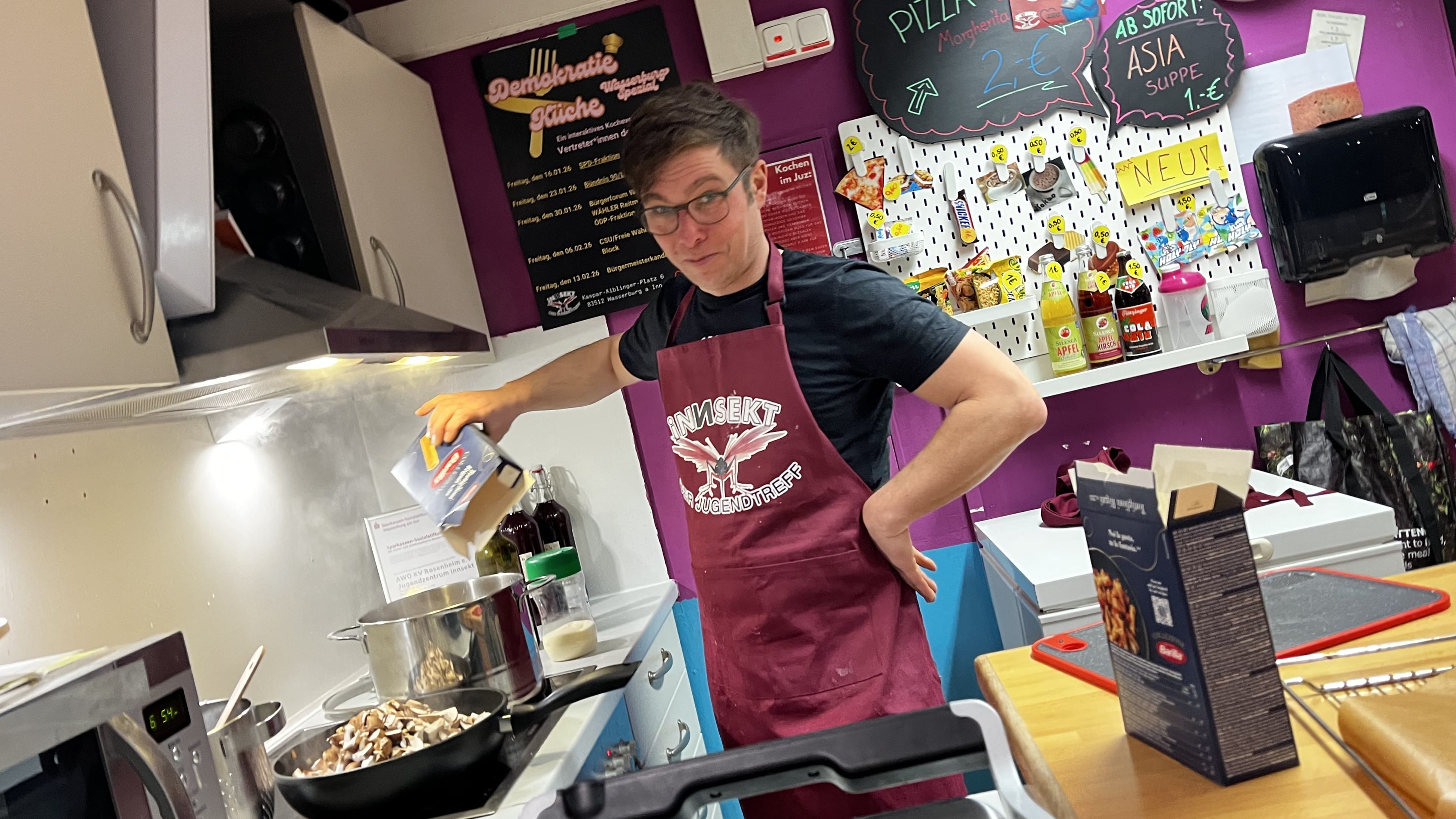 Foto: ein junger Erwachsener mit einer Kochschürze steht in einer Küche und schüttet Nudeln in einen Topf mit kochendem Wasser. Auf der Schürze ist das Logo des iNNSEKTs zu sehen und im Hintergrund das Plakat mit den Terminen der Demokratie-Küche.