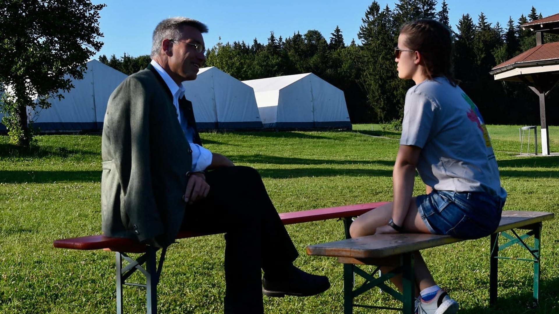 Der Landrat interviewt Hannah. Beide sitzen sich auf Bierbänken gegenüber. Im Hintergrund sieht man große weiße Zelte. Hannah trägt das aROund T-Shirt.