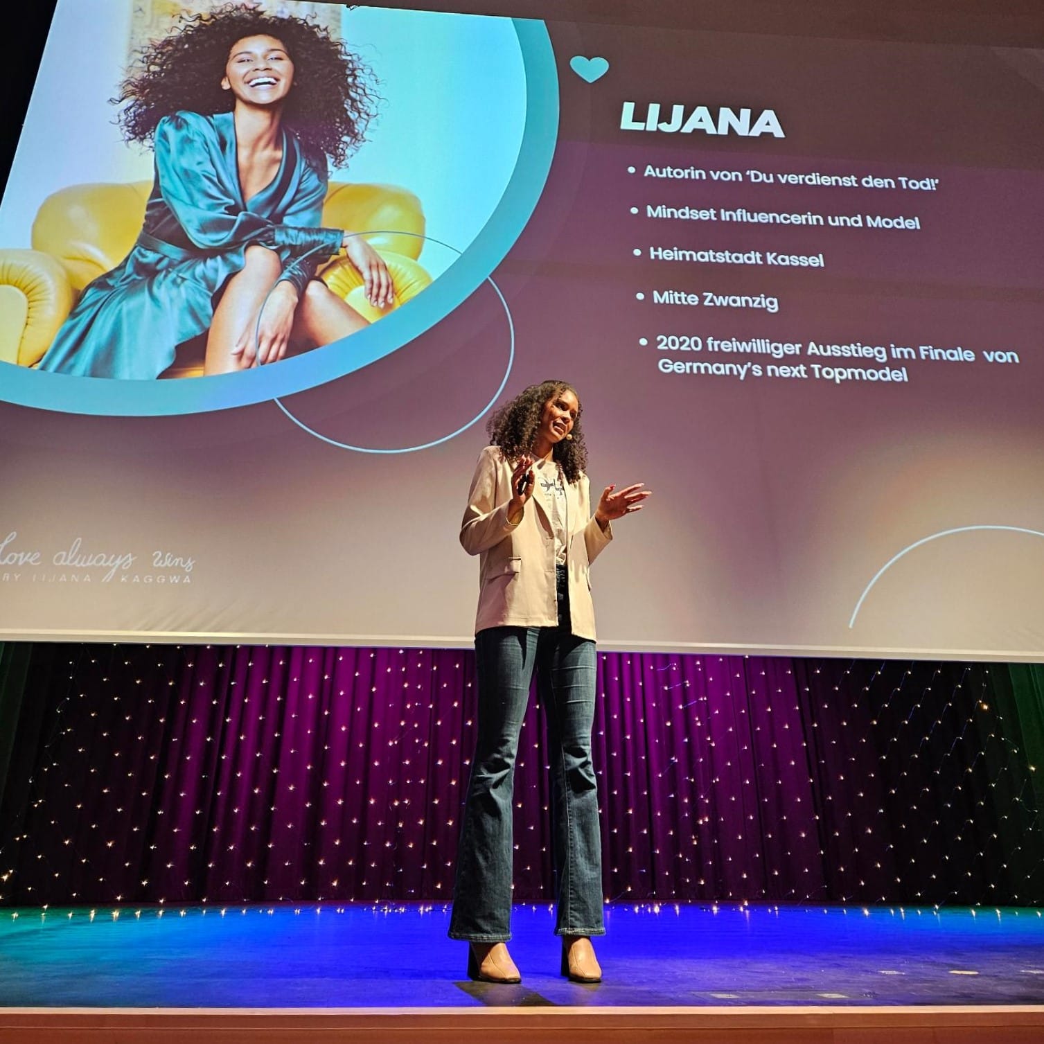 Foto: Lijana steht auf der Bühne vor einer Leinwand, auf die ein paar Schlagworte zu ihrer Person projiziert sind, die alle im Beitrag genannt werden