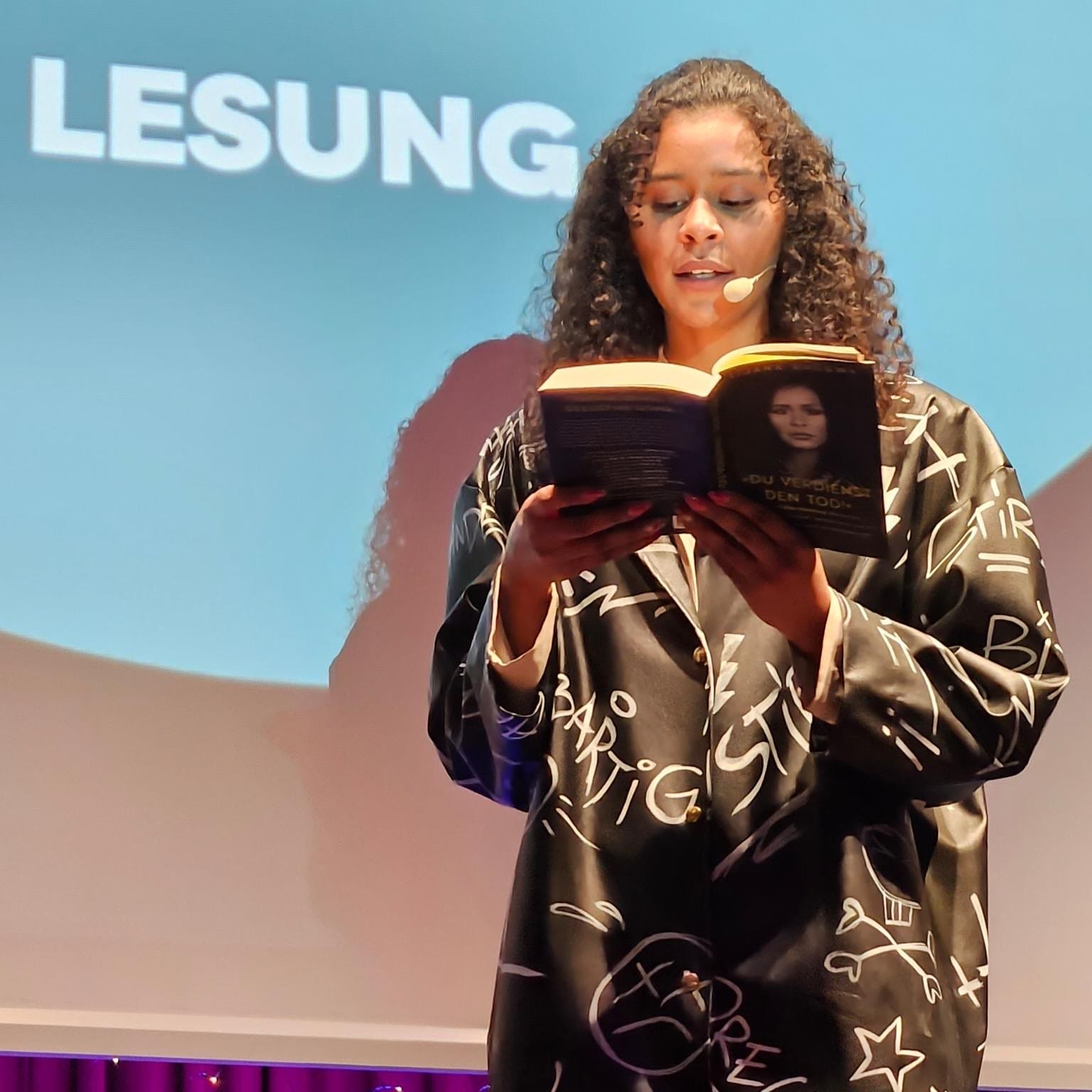Foto: Lijana Halbnah mit ihrem Buch in der Hand, aus dem sie zitiert. Sie trägt einen Mantel auf den Hassbotschaften geschrieben sind