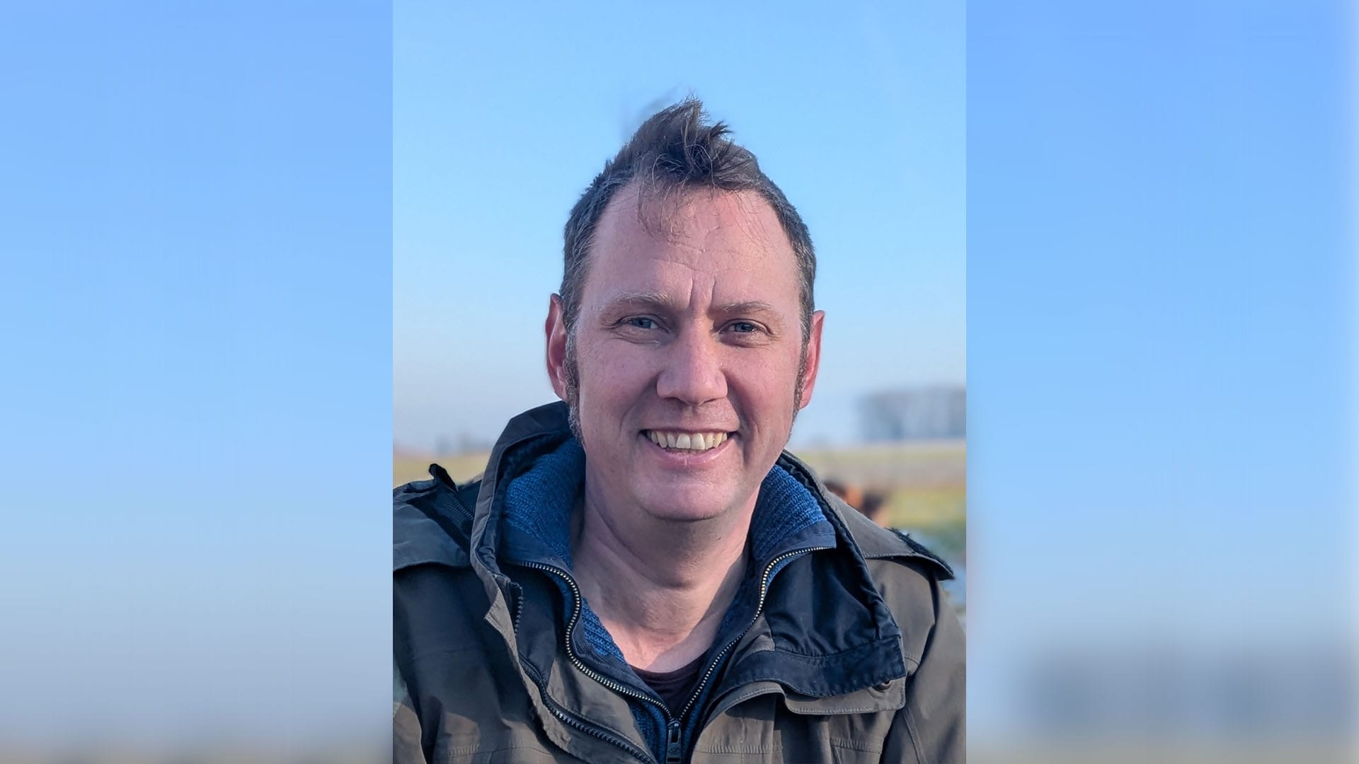 Portrait: Ein Mann mit Parka steht vor einem Feld und lächelt in die Kamera