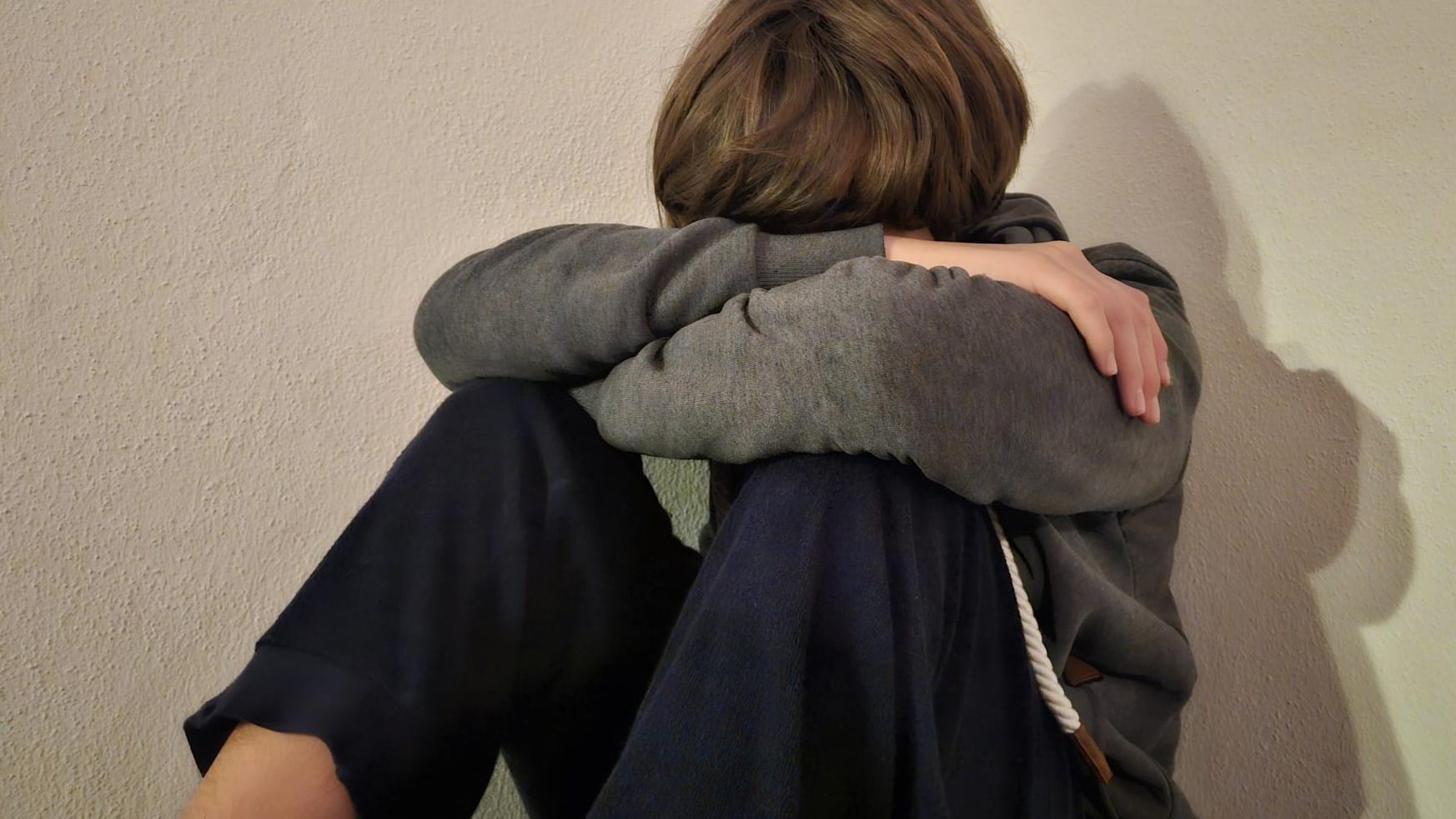 Foto von einem Teenager, der an einer kahlen Wand sitzt und traurig den Kopf auf die verschränkten Arme gelegt hat.