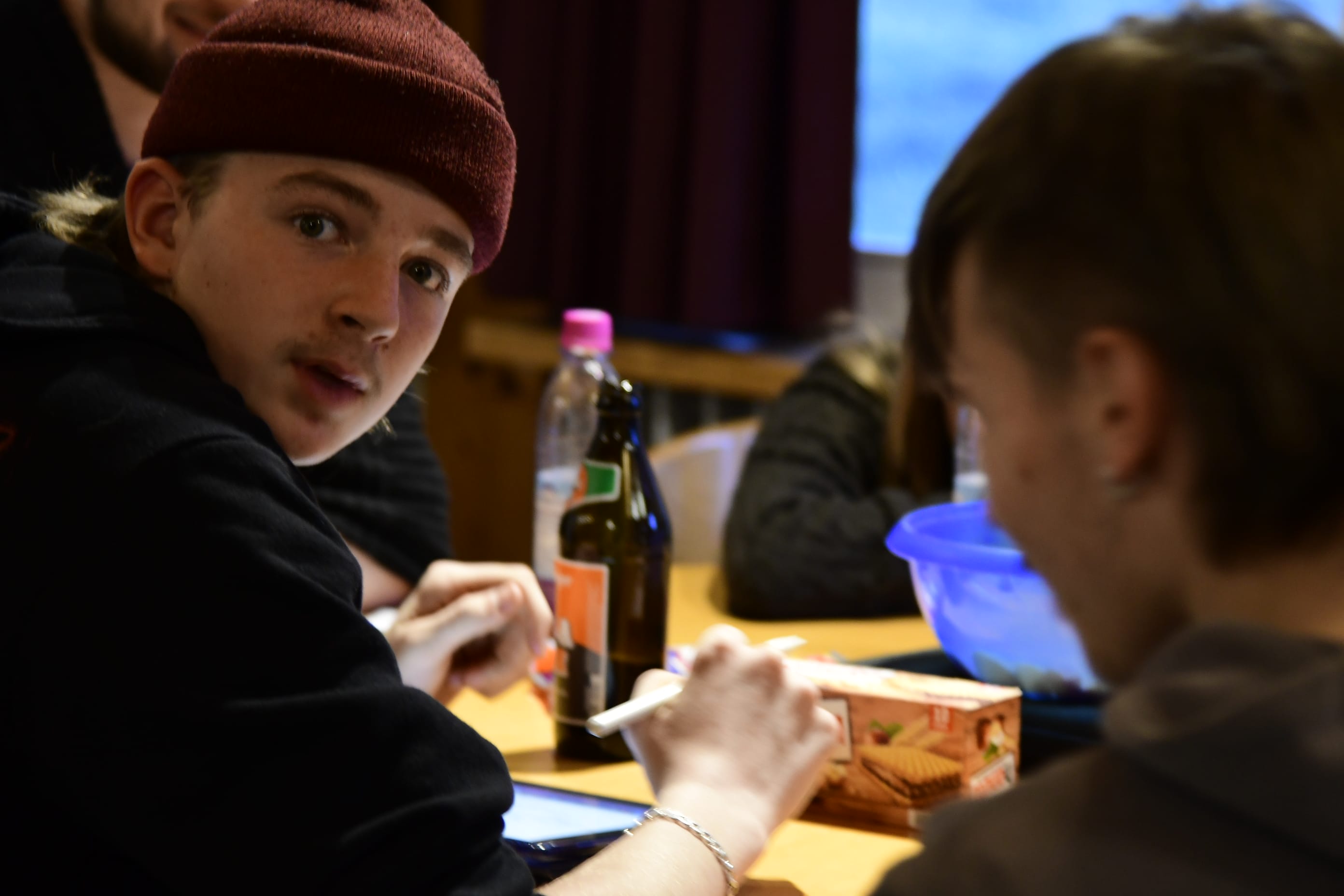 Foto: Ein junger Mann schaut über seine Schulter direkt in die Kamera. Er sitzt mit anderen an einem Tisch.