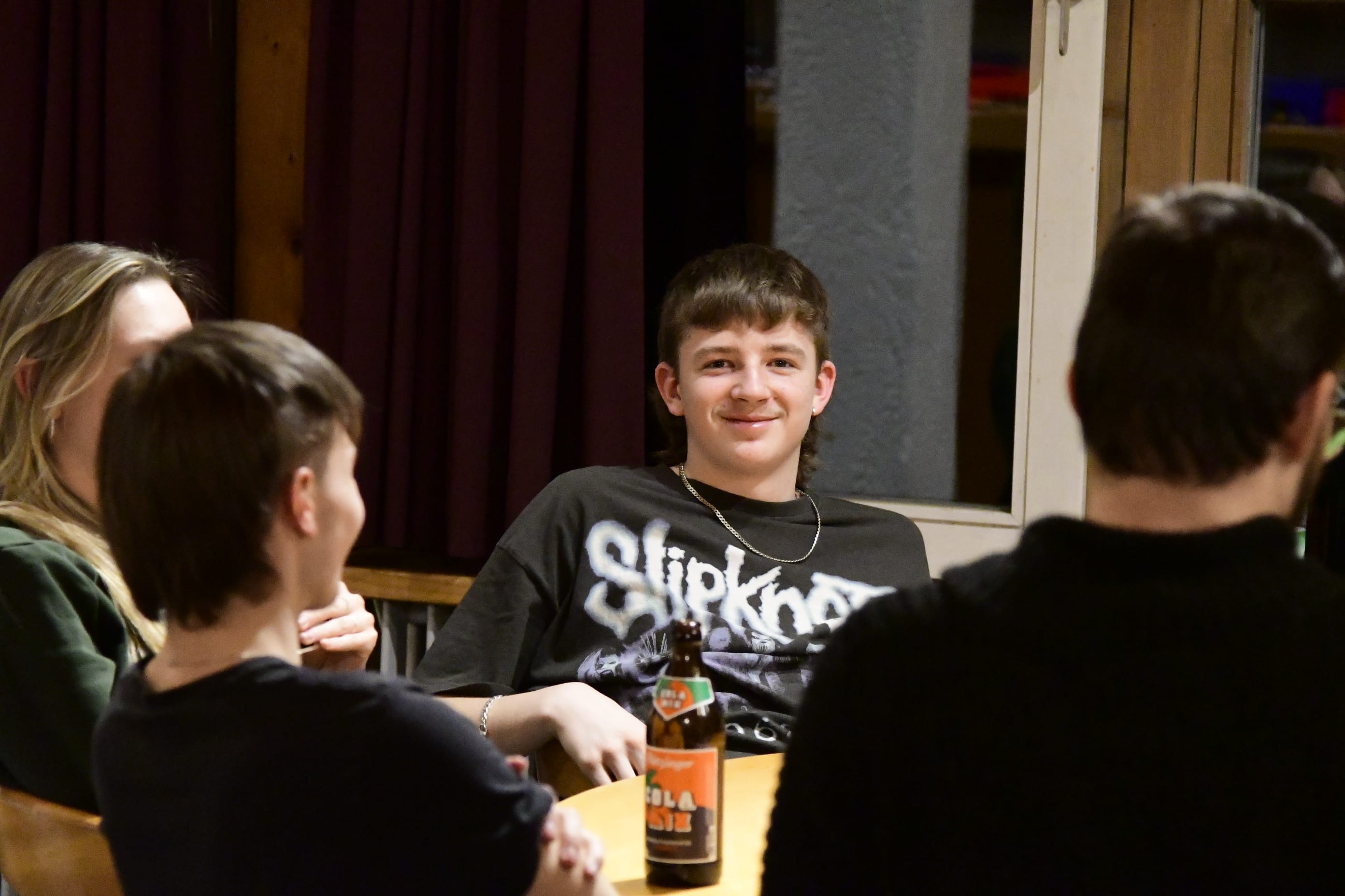 Foto: Ein junger Mann mit einer Mütze schaut über seine Schulter in die Kamera. Er sitzt mit anderen an einem Tisch.