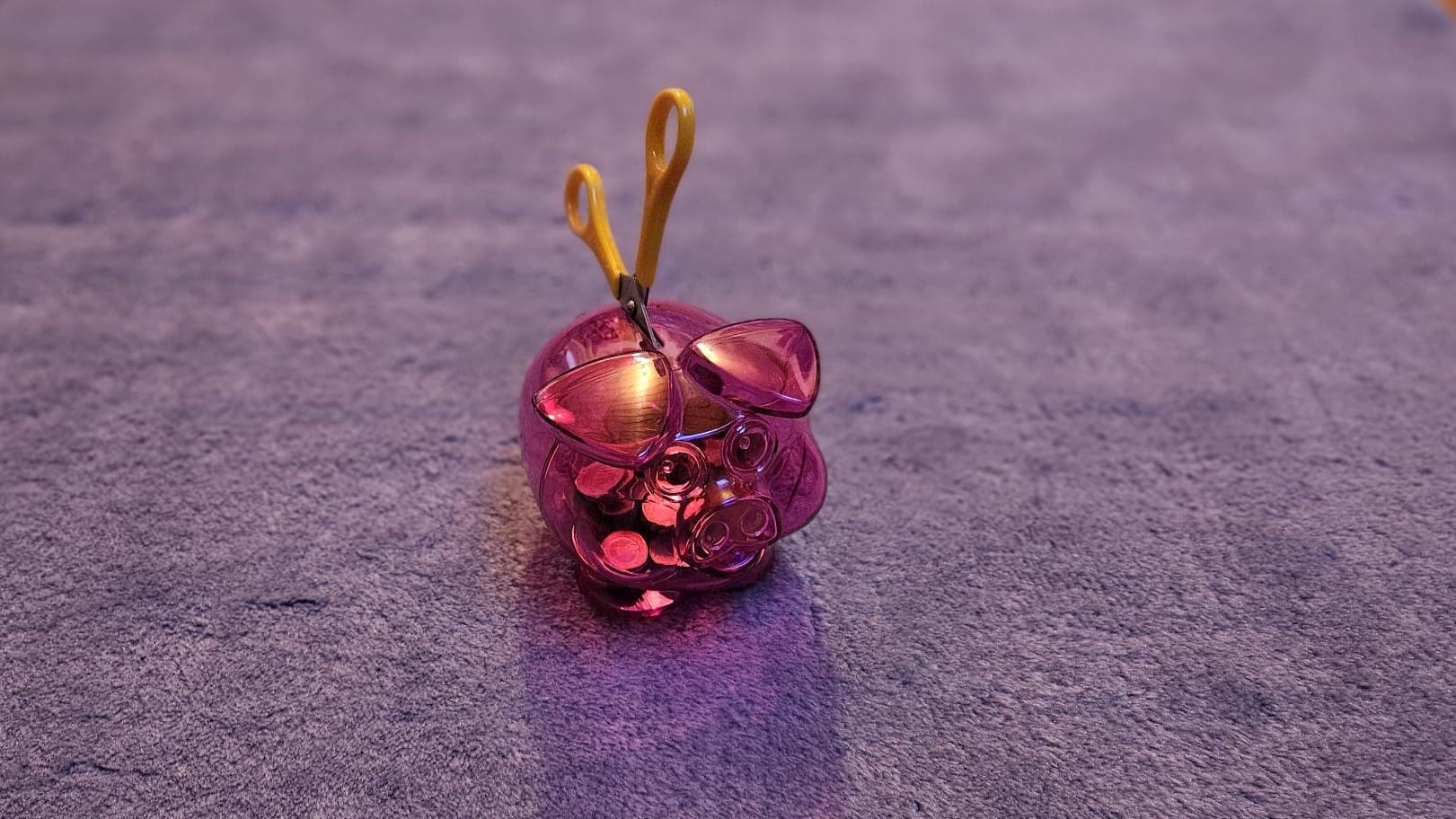 Foto: Ein mit Euro- und Cent-Münzen gefülltes, transparent-rosa Sparschwein auf einem Teppich. Im Schlitz oben steckt eine Schere.