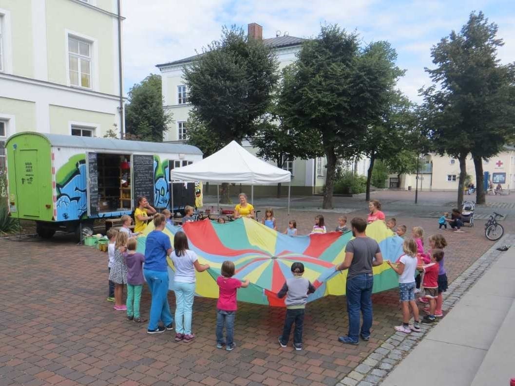 Foto: Kinder und Betreuer stehen im Kreis und halten ein Schwungtuch in Regenbogenfarben
