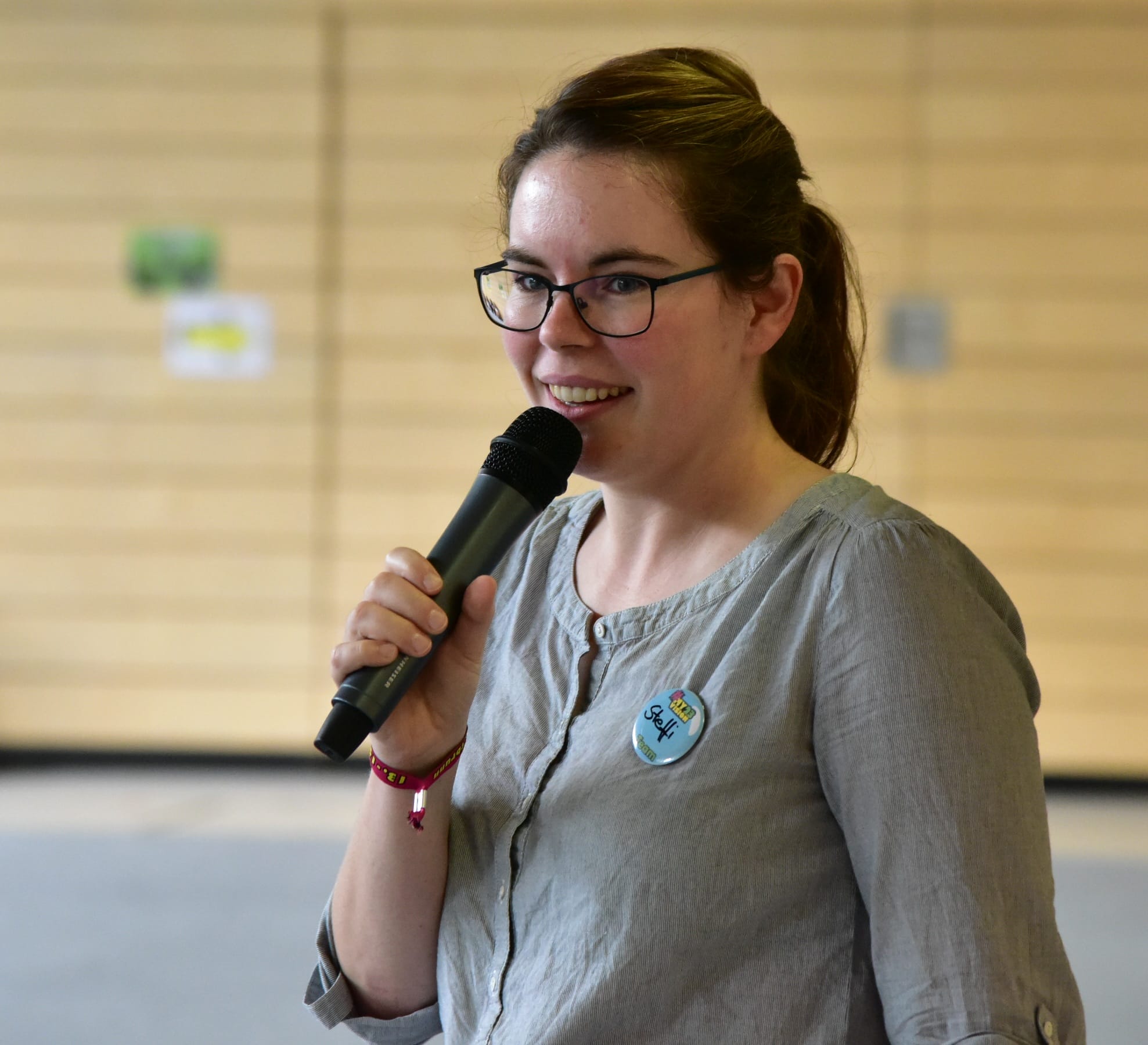 Foto: Eine junge Frau in einer Turnhalle spricht in ein Handmikrofon. Im Hintergrund hängen Plakate an der Wand.