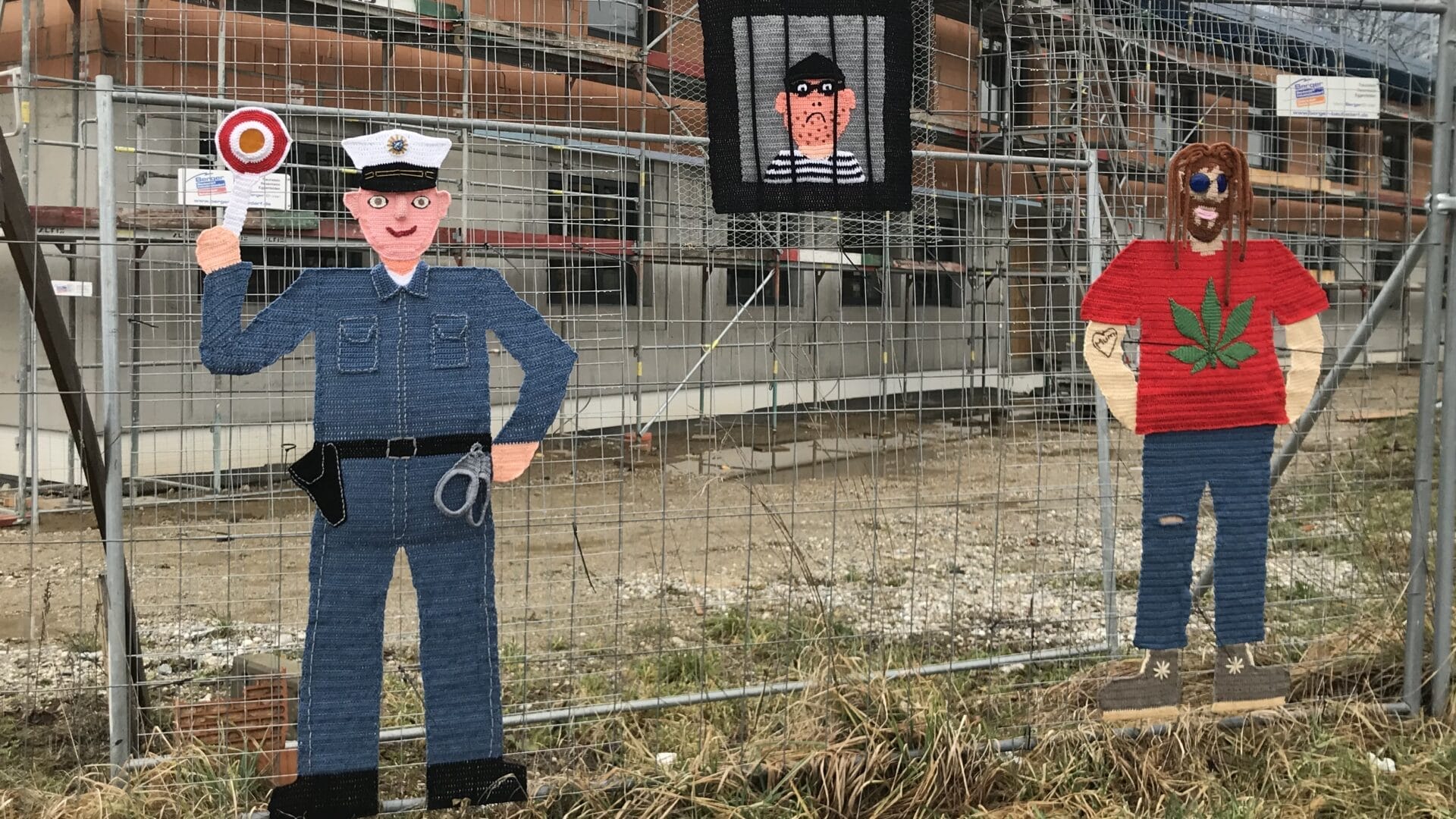 Foto: An einem Bauzaun hängen aus Wolle gestrickte Figuren eines Polizist, eines Gefangenen und eines Mannes mit Rastahaaren und Canabis Pflanze auf dem Pulli