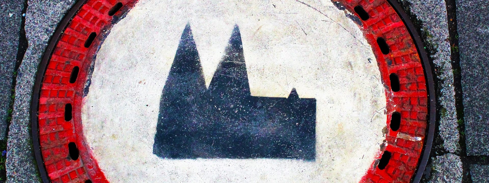 Foto von einem Gullideckel, aud dem der Kölner Dom aufgemalt ist. Erinnert an ein Einfahrt-Verboten Schild mit einem Roten Ring und einer weißen Innenfarbe.