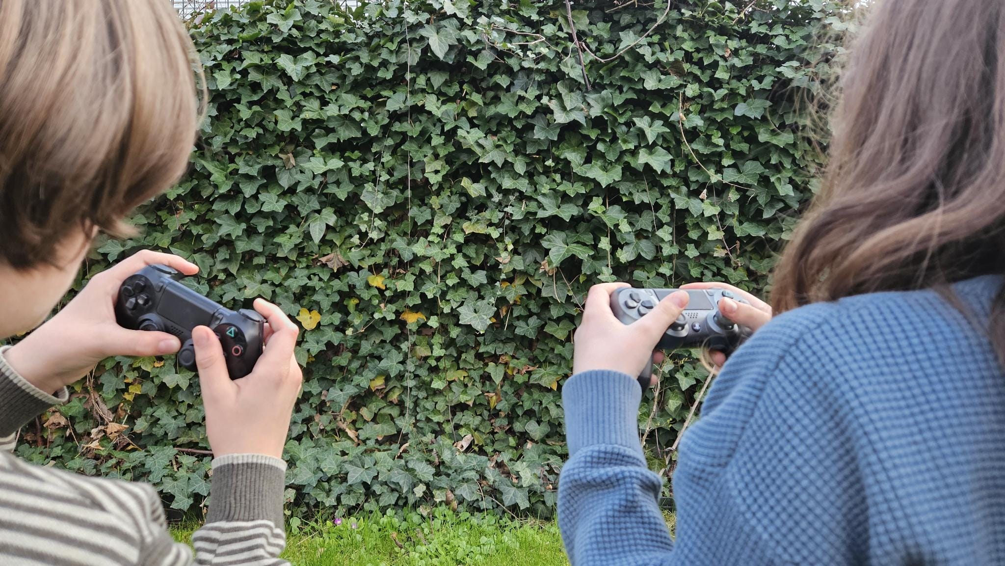 Foto: Zwei Jugendliche mit Gaming-Controllern sitzen outdoor vor einer grün mit Weinlaub bewachsenen Wand