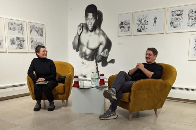 Foto: Auf zwei Sesseln sitzen eine Frau und ein jüngerer Mann mit Brille und Turnschuhen. Auf der Wand im Hintergrund ist überlebensgroß ein Boxer abgebildet.