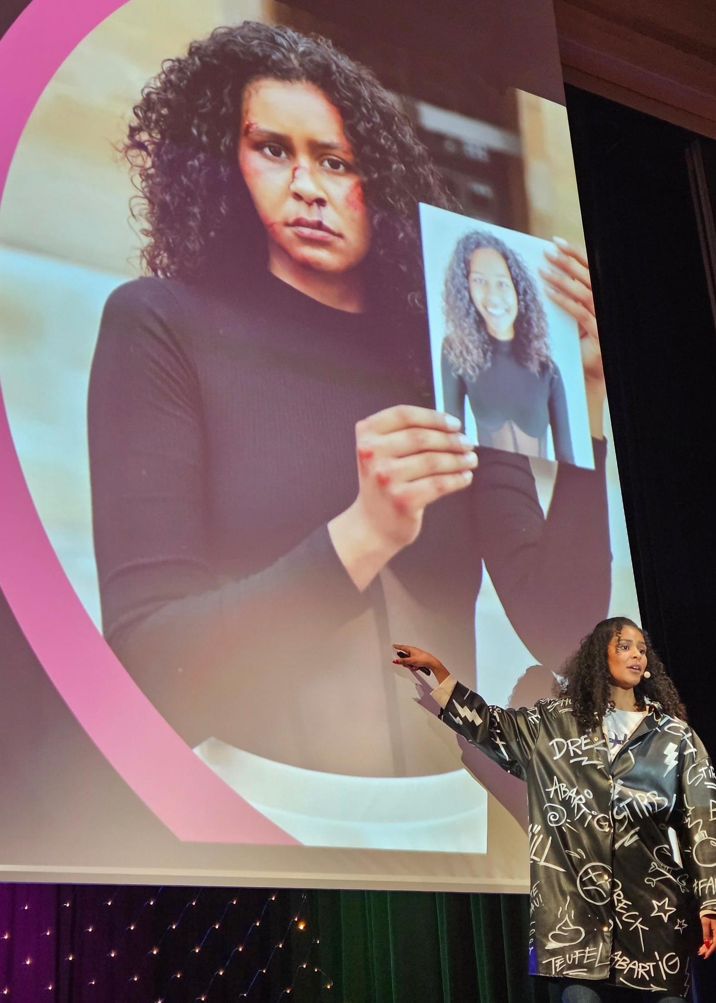 Foto: Lijana zeigt auf die Leinwand hinter ihr auf ein Bild von ihr mit aufgeschminkten Blessuren